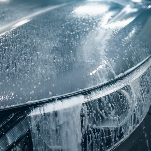 Car Wash, active cleaning foam of first phase flows down hood of auto and headlight, close up.
